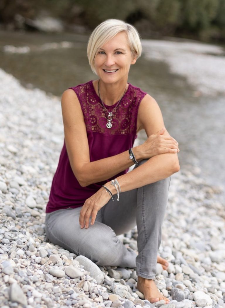Frau mit kurzen blonden Haaren, lächelnd, sitzt auf Steinen am Ufer.