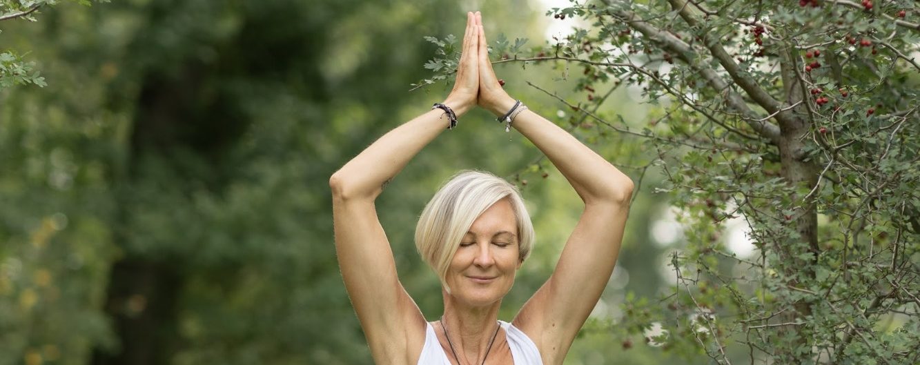 Ältere Frau in Yogapose im Freien, mit offenem Lächeln und erhobenen Händen.