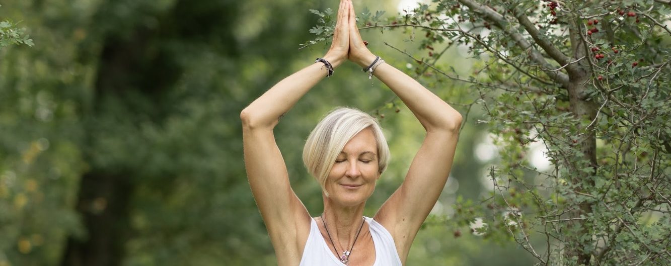 Spirituelle Coaching, Emtspanungsmassage Ältere Frau in Yogapose im Freien, mit offenem Lächeln und erhobenen Händen.